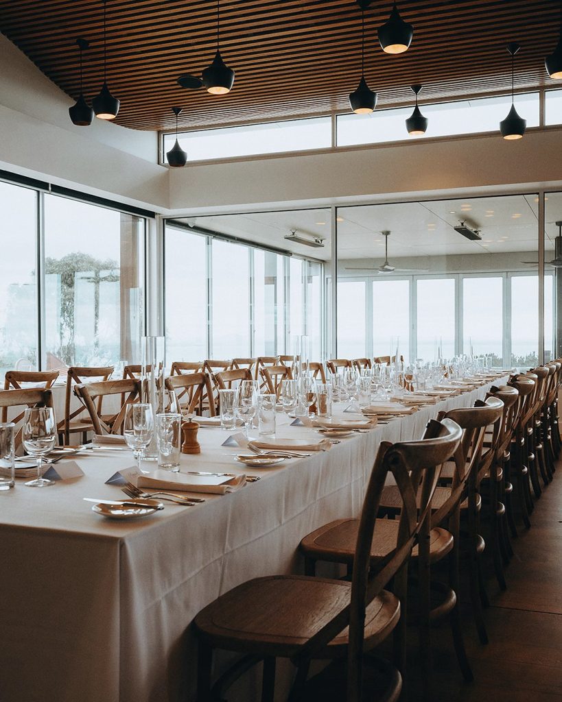 Long banquet table in cream linen running the length of a venue under black pendant lights