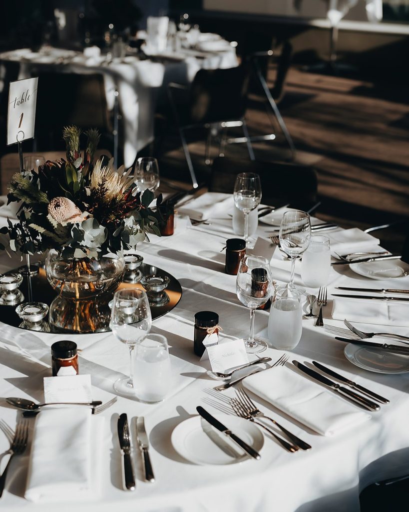 Banquet table set in white linen under low light, with a native protea and banksia centrepiece