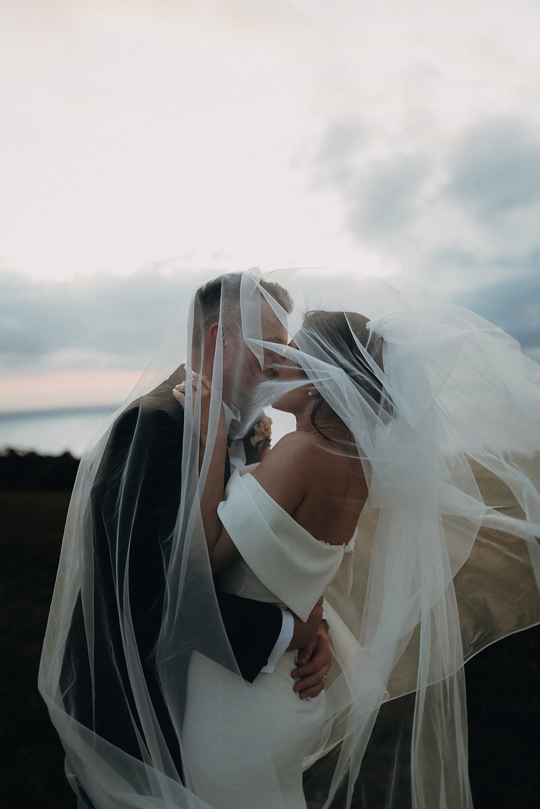 Bride and groom kissing beneath a wind-caught veil at dusk, ocean behind