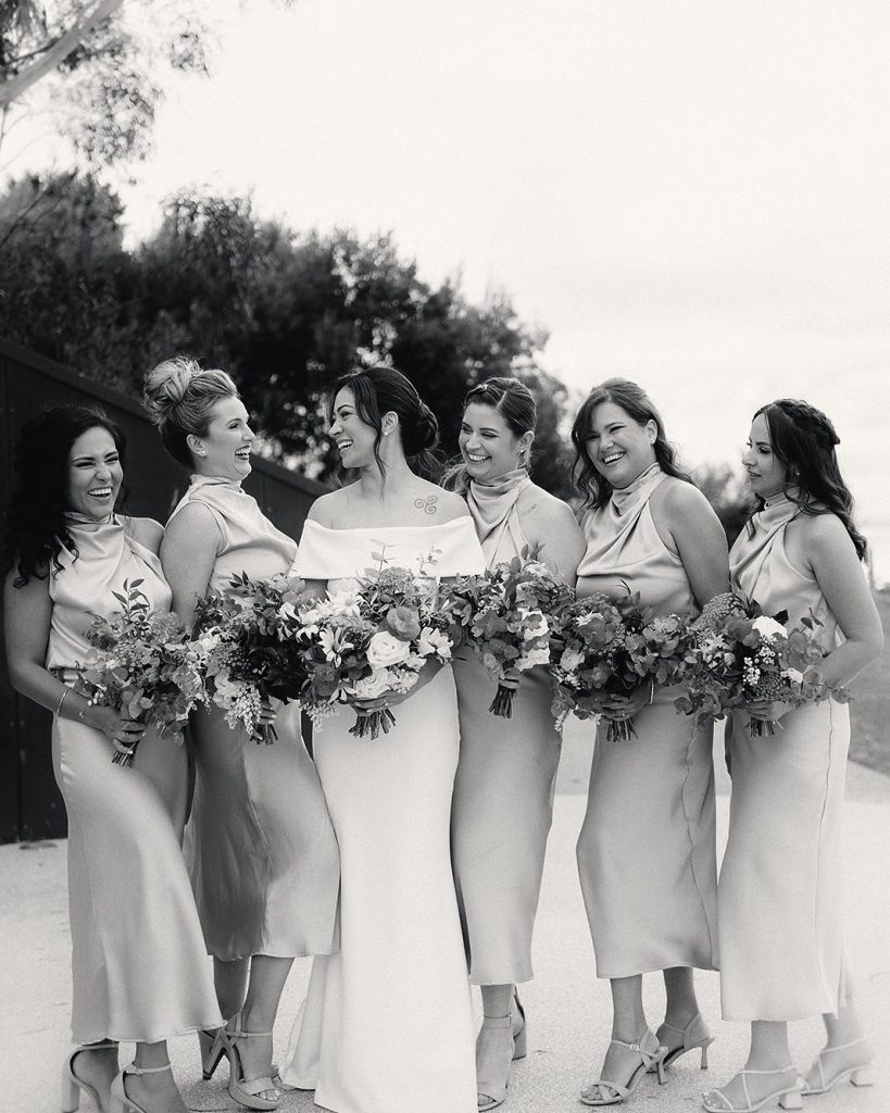 Bride and five bridesmaids in champagne satin laughing together, black and white