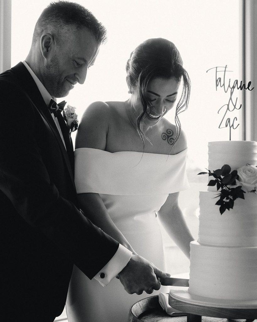 Bride and groom cutting a three-tier cake by a bright window, black and white