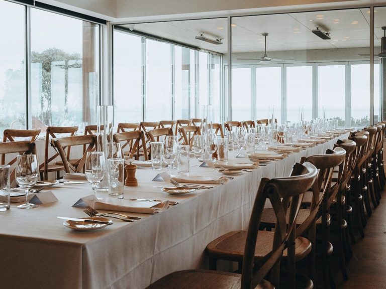 Long banquet table set in cream linen under pendant lights, ocean through the glass